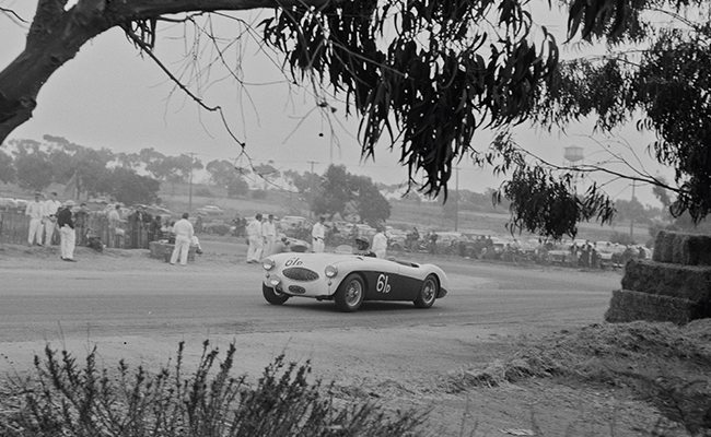 A vintage black and white photograph captures a convertible sports car, racing at the Torrey Pines Road Races in the 1950s, with spectators and eucalyptus trees in the background.