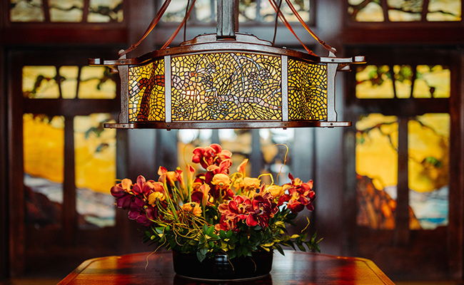 An Arts and Crafts stained glass lantern hangs in the foreground over a floral centerpiece, with the Judson Studios-crafted front doors of The Lodge at Torrey Pines visible in the background.